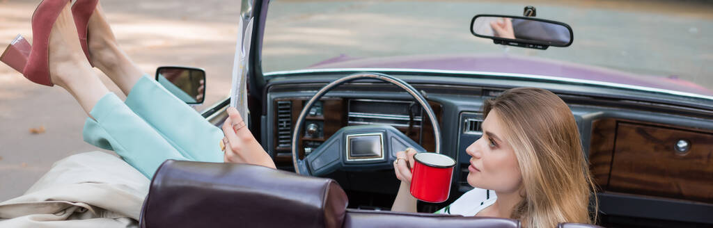 молодая женщина, держащая чашку кофе и глядя на карту, сидя в кабриолете с поднятыми ногами на двери, баннер - Фото, изображение