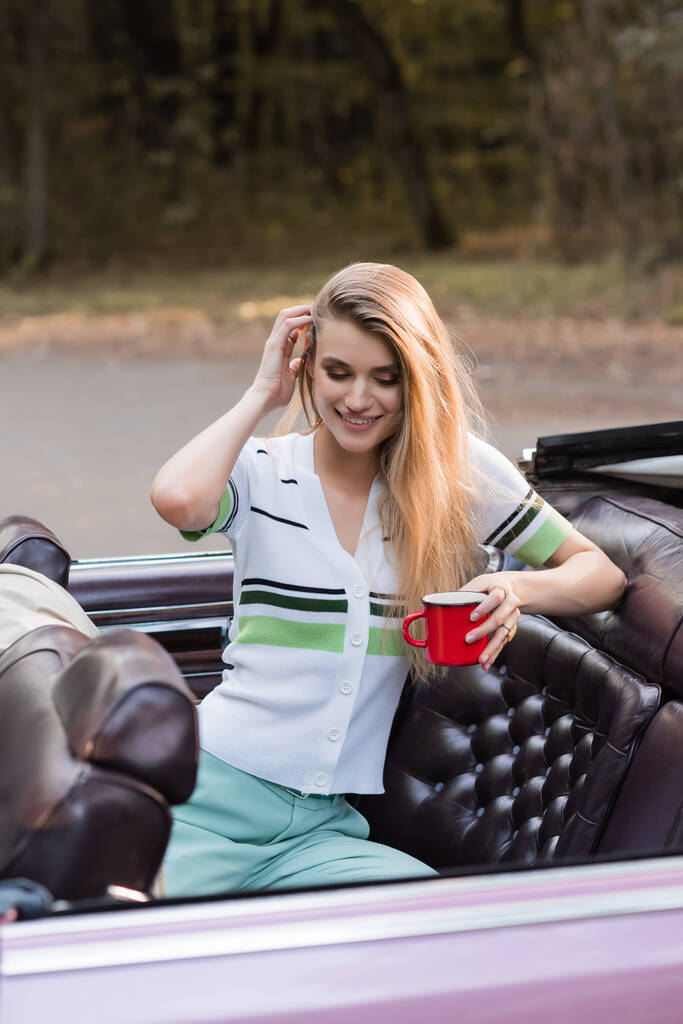 mujer sonriente tocando el cabello mientras sostiene la taza de café en cabriolet vintage - Foto, Imagen