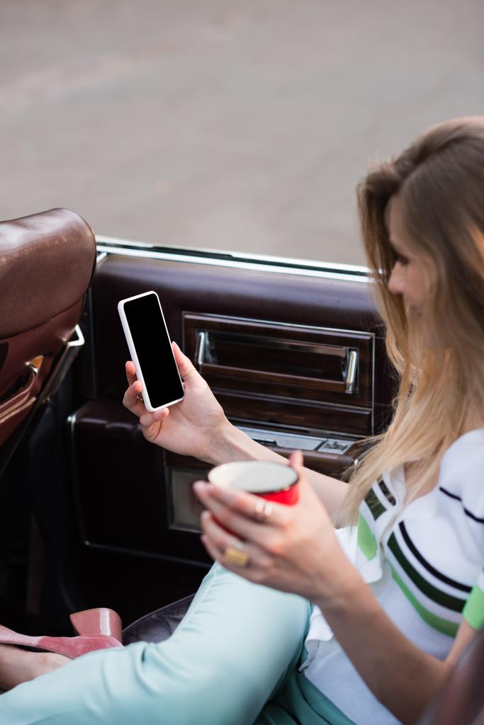 Önplanda bulanık bir şekilde Cabriolet 'te otururken elinde beyaz ekran ve bir fincan kahve olan akıllı telefonu tutan genç bir kadın. - Fotoğraf, Görsel