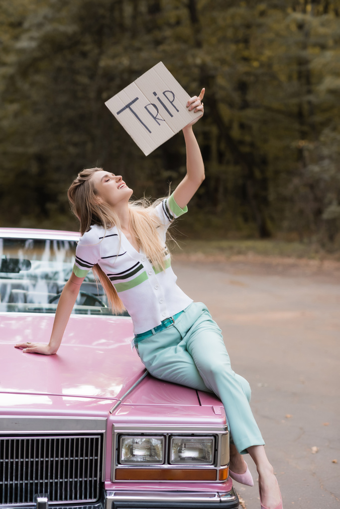 donna sorridente che tiene cartello con scritte di viaggio mentre si siede sul cappuccio di cabriolet - Foto, immagini
