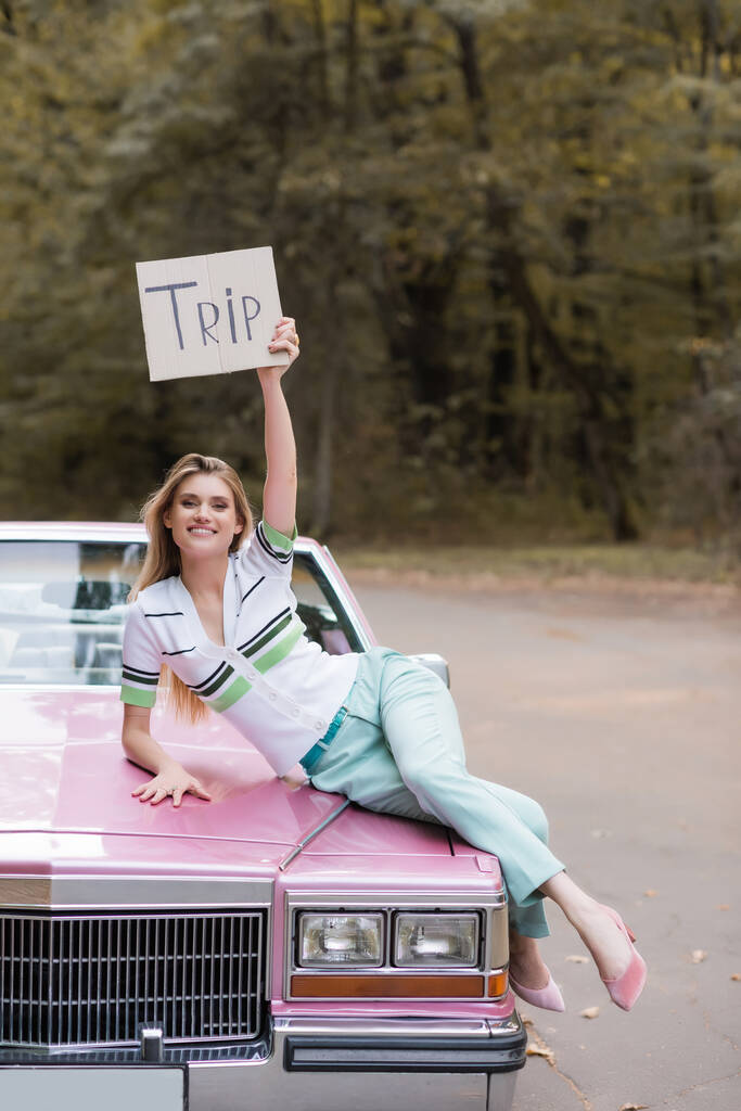 stylowa młoda kobieta trzymająca plakietkę z literami podróży leżąc na kabriolecie  - Zdjęcie, obraz