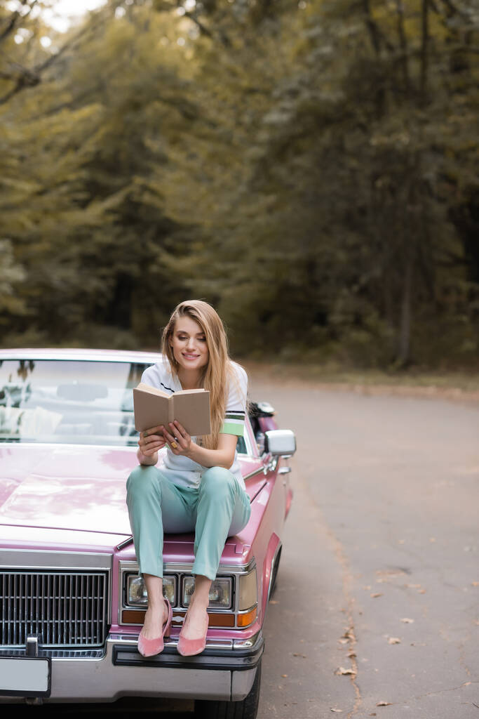 χαμογελαστή γυναίκα κάθεται στο καπό του cabriolet και διαβάζοντας το βιβλίο - Φωτογραφία, εικόνα