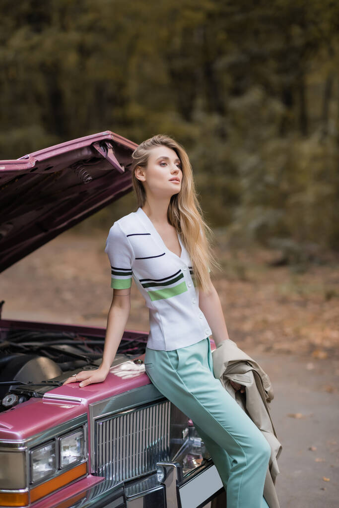 mujer elegante joven mirando hacia otro lado mientras que de pie cerca de coche retro roto con capucha abierta - Foto, Imagen