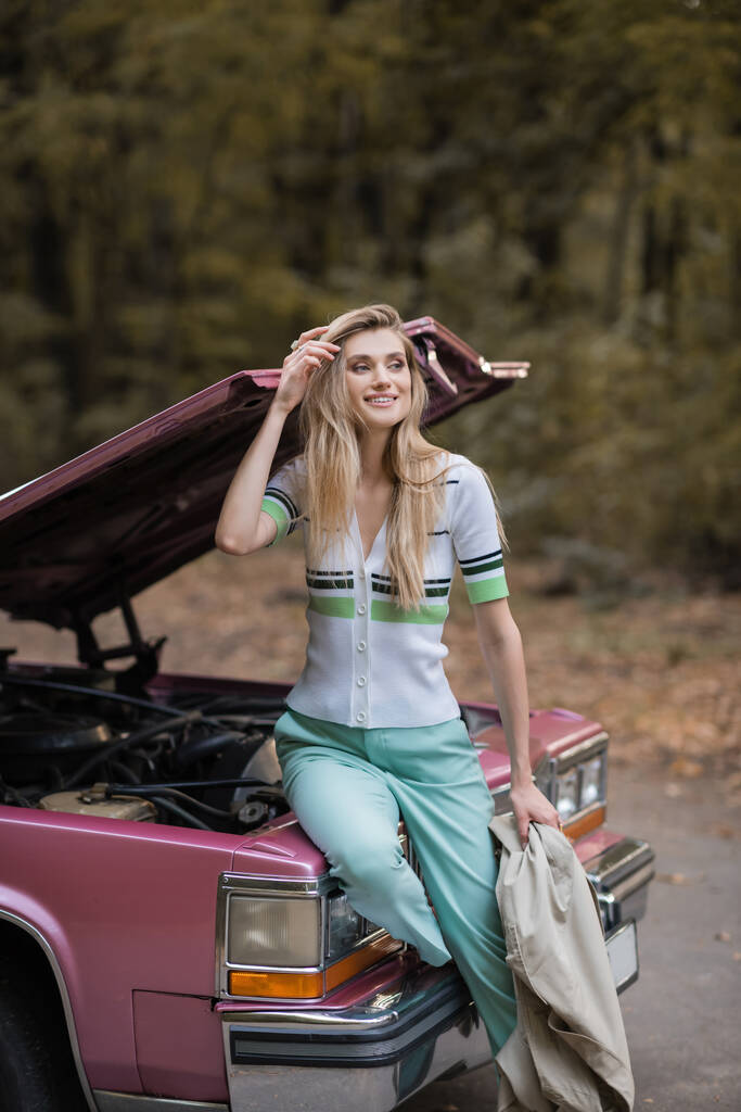 улыбающаяся женщина касается волос и смотрит в сторону, сидя на сломанной машине с открытым капотом - Фото, изображение