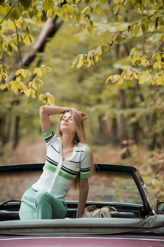 jeune femme élégante tenant la main au-dessus de la tête tout en posant dans le cabriolet avec les yeux fermés - Photo, image
