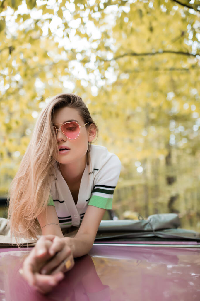 молодая стильная женщина в солнечных очках, опирающаяся на кабриолет на размытом переднем плане - Фото, изображение