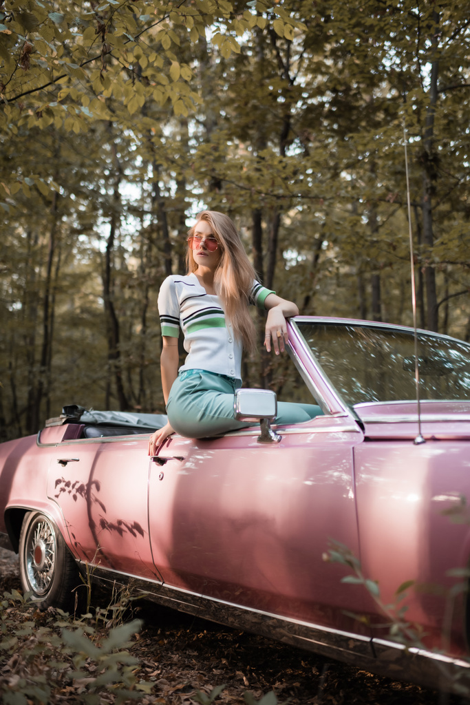 stijlvolle vrouw in een zonnebril weg te kijken terwijl zitten op de deur van converteerbare auto - Foto, afbeelding