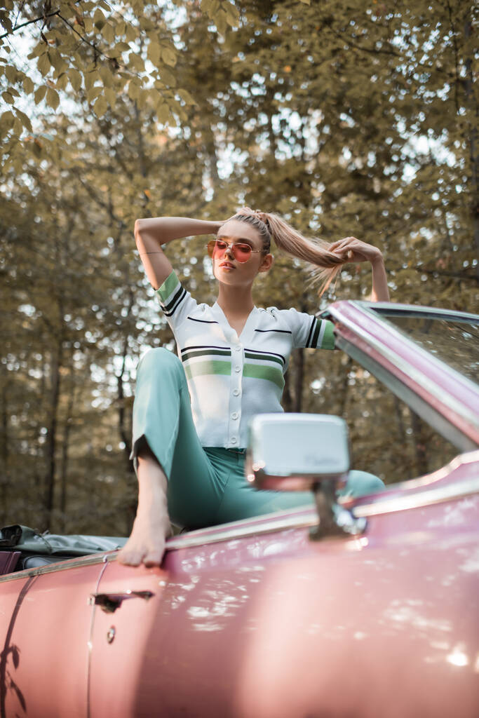 стильная босиком женщина в солнечных очках трогает волосы, позируя в кабриолете на размытом переднем плане - Фото, изображение