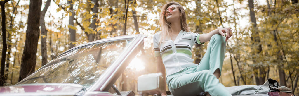 stijlvolle vrouw in een zonnebril weg te kijken terwijl poseren in retro converteerbare auto in de zon, banner - Foto, afbeelding