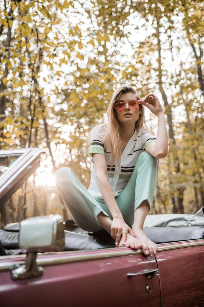 stylová bosá žena dotýkající se slunečních brýlí při pózování v kabrioletu v lese na rozmazaném popředí - Fotografie, Obrázek