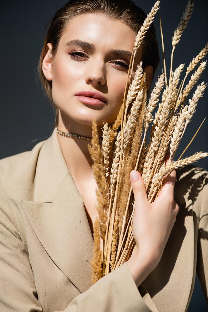 young woman in beige blazer looking at camera near wheat spikelets on dark grey - Photo, Image
