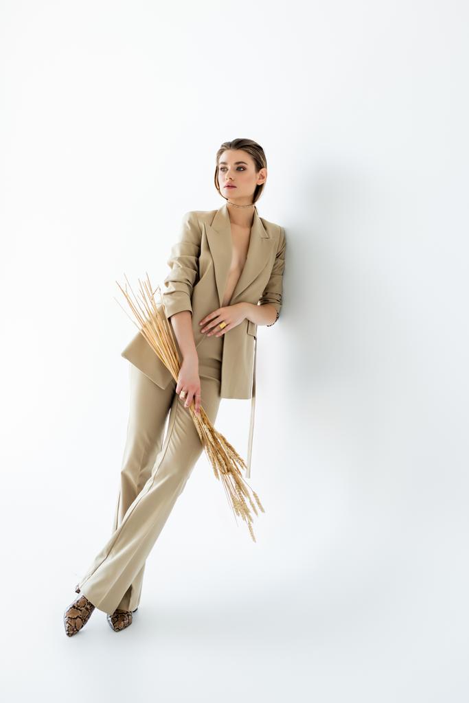 повна довжина молодої жінки в бежевому формальному одязі позує, тримаючи пшеницю на білому
 - Фото, зображення