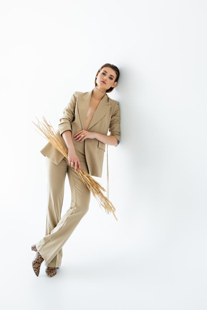 πλήρες μήκος της νεαρής γυναίκας σε μπεζ κοστούμι που θέτει, ενώ κρατώντας σιτάρι σε λευκό - Φωτογραφία, εικόνα