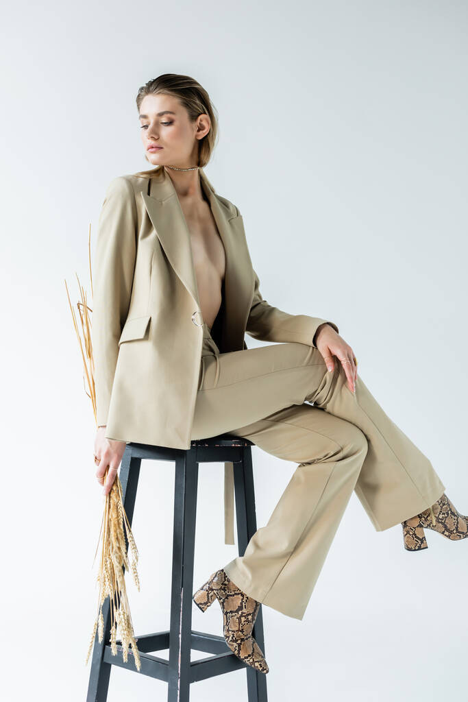sexy mladý model v obleku sedí na stoličce a drží pšeničné hřeby na bílém - Fotografie, Obrázek