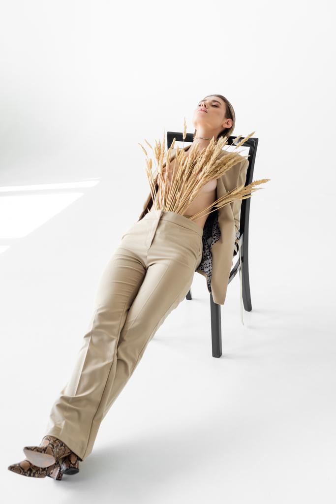 stilvolles Modell in beige Anzug mit Weizen posiert auf Stuhl auf weißem Hintergrund - Foto, Bild