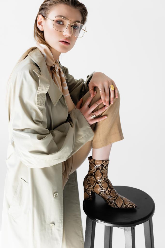 jovem na moda em casaco de trincheira e cachecol posando perto da cadeira no branco - Foto, Imagem