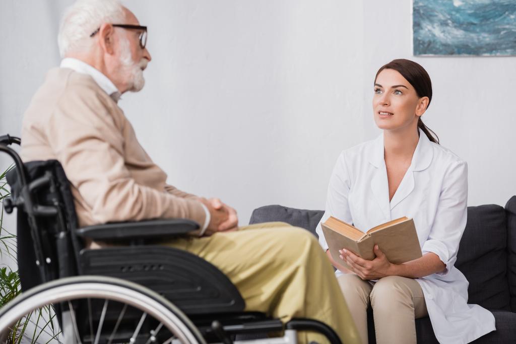 sociaal werker in witte jas lezen boek voor oudere gehandicapte man in een rolstoel op wazig voorgrond - Foto, afbeelding