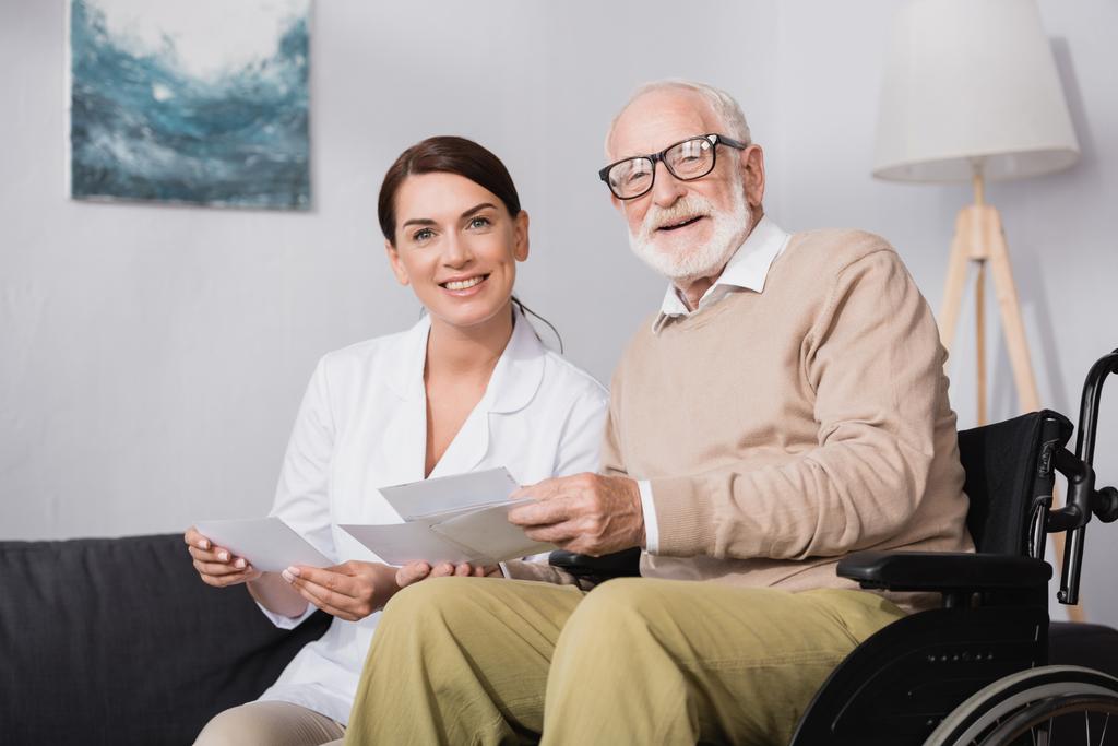 smiling social worker and aged man looking at camera while holding photos - Photo, Image