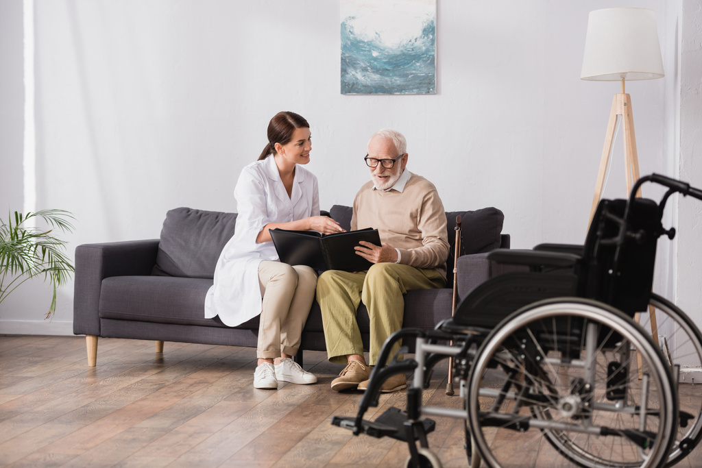 geriatrická zdravotní sestra a starý muž prohlížení fotoalbum v blízkosti invalidního vozíku na rozmazané popředí - Fotografie, Obrázek