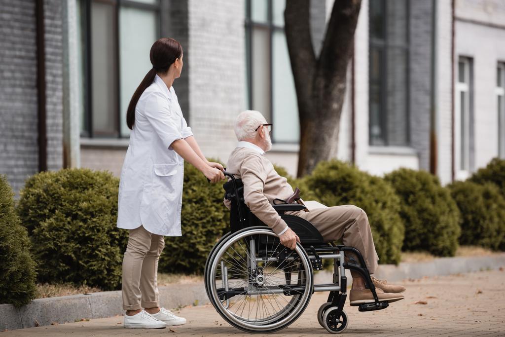 sociaal werker wandelen met bejaarde gehandicapte man op rolstoel buiten - Foto, afbeelding