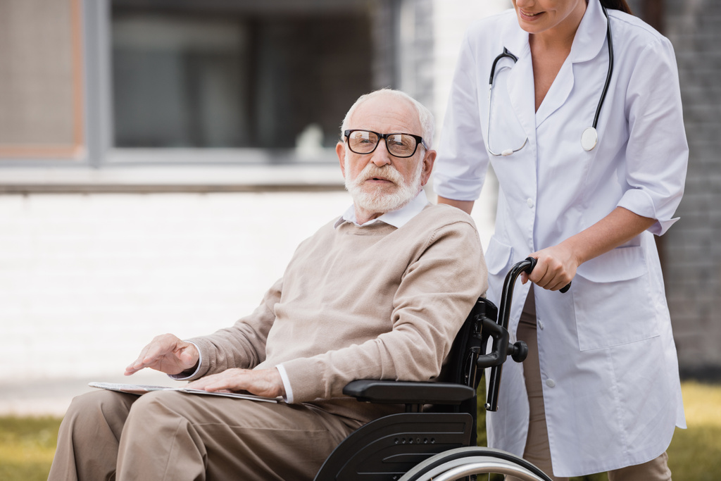 starý zdravotně postižený muž na vozíku při pohledu na kameru při chůzi venku s geriatrickou zdravotní sestrou - Fotografie, Obrázek