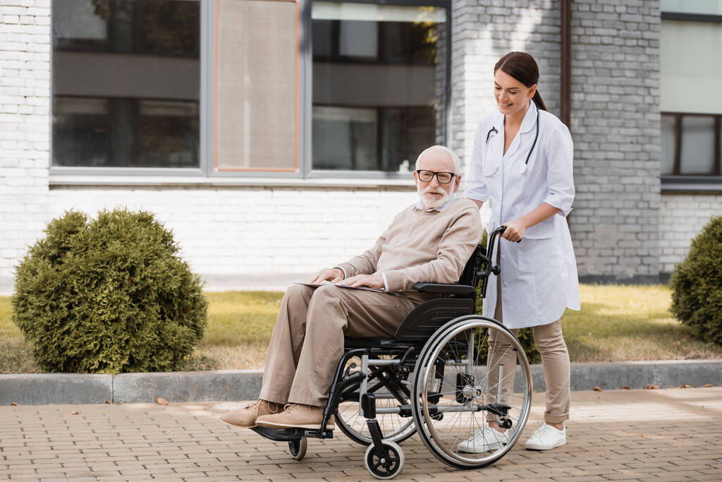 sonriente trabajador social caminando con hombre discapacitado en silla de ruedas al aire libre - Foto, imagen