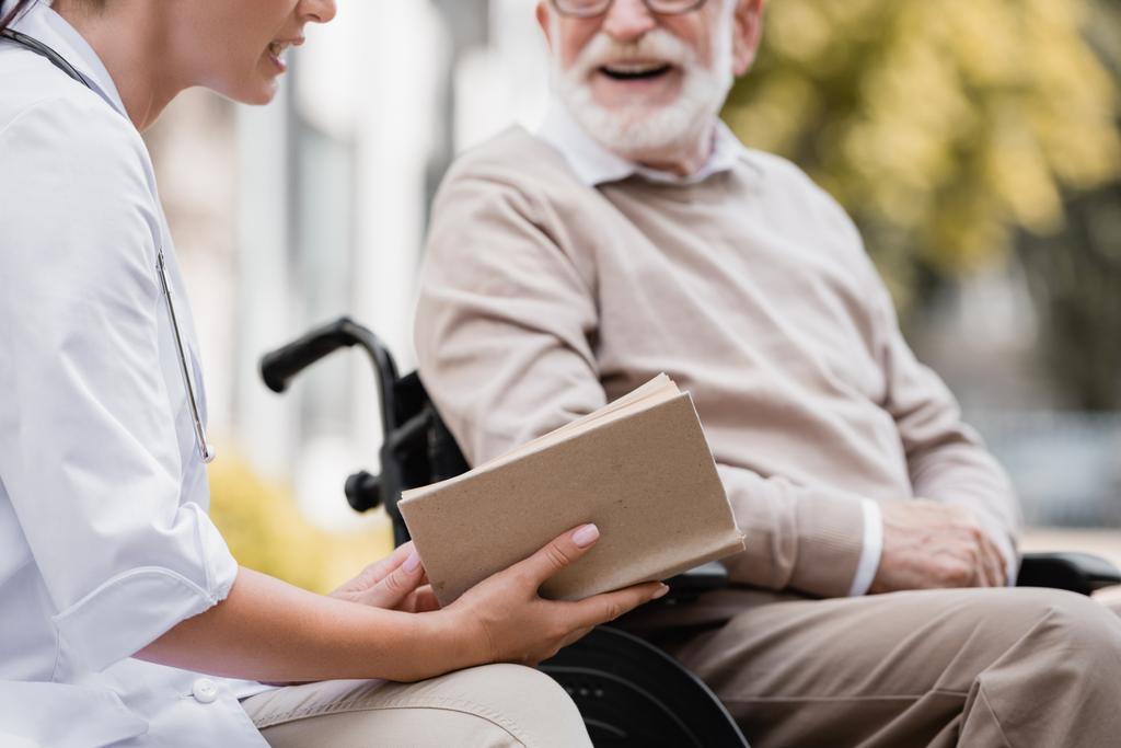 bijgesneden weergave van geriatrische verpleegkundige lezen boek aan oudere man op wazige achtergrond - Foto, afbeelding