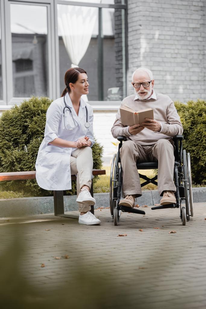 Behinderter älterer Mann liest im Rollstuhl neben Sozialarbeiterin auf Bank im verschwommenen Vordergrund - Foto, Bild