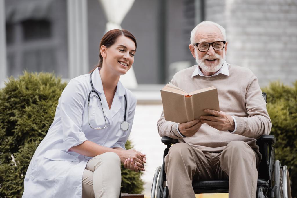 весела геріатрична медсестра біля літнього чоловіка з обмеженими можливостями читає книгу в інвалідному візку на відкритому повітрі
 - Фото, зображення