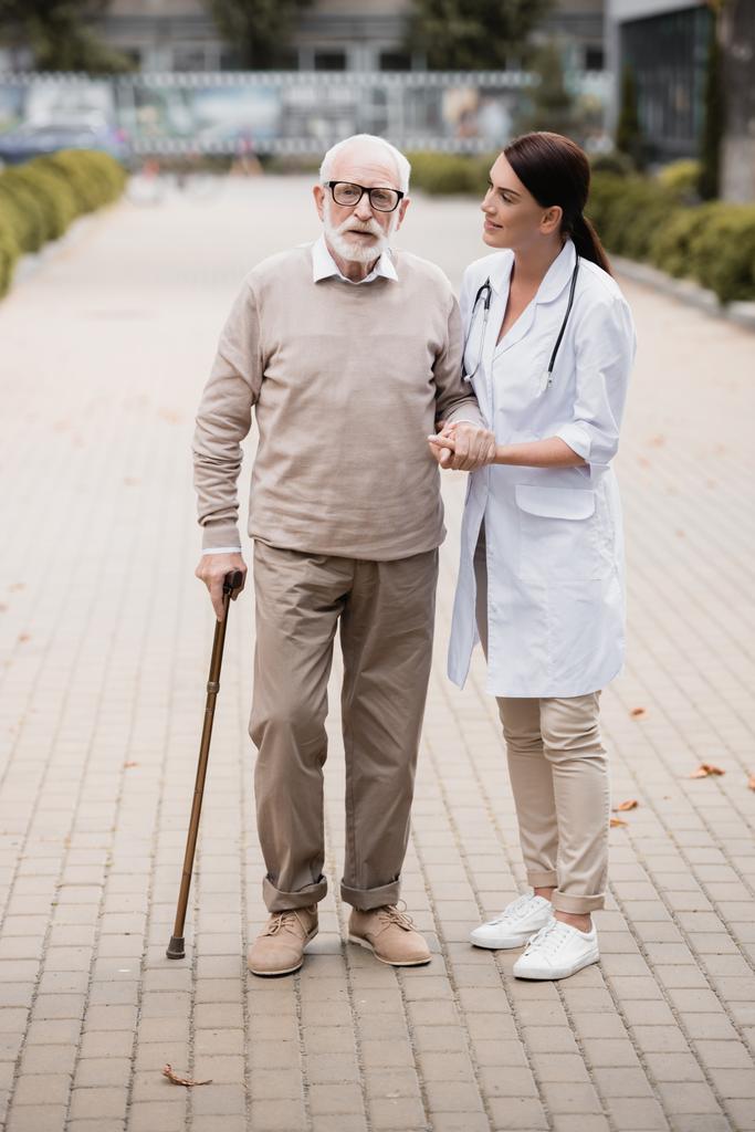 sociaal werker ondersteuning van oude man met wandelstok terwijl wandelen samen - Foto, afbeelding