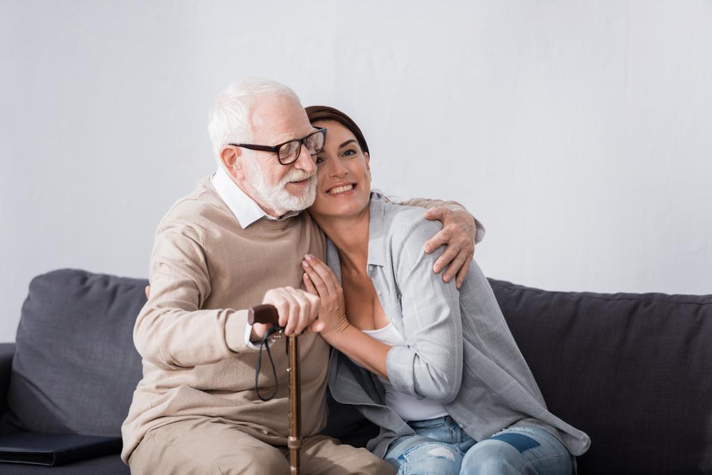 ευγνώμων ηλικιωμένος άνδρας αγκαλιάζει γηριατρική νοσοκόμα, ενώ κάθεται στον καναπέ στο σπίτι - Φωτογραφία, εικόνα