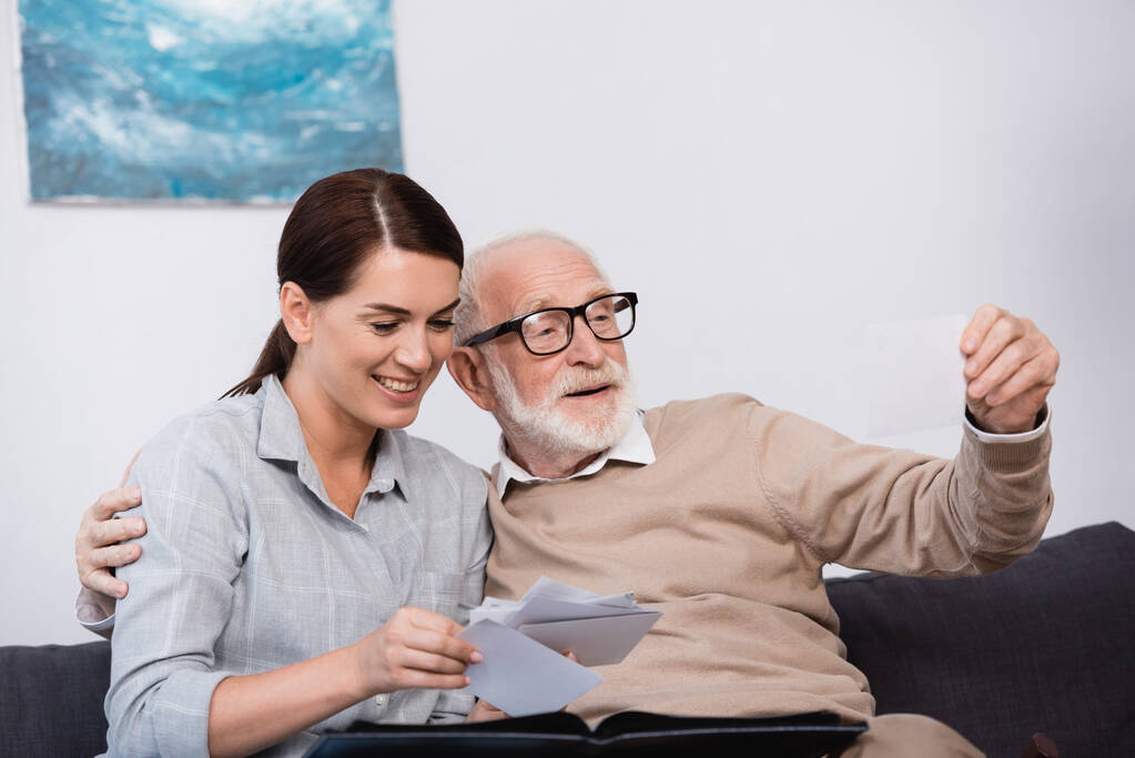 mujer sonriente con padre anciano mirando fotos familiares juntos en casa - Foto, imagen