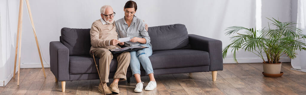 ενήλικη γυναίκα και ηλικιωμένος πατέρας κοιτάζοντας οικογενειακές φωτογραφίες, ενώ κάθεται στον καναπέ στο σπίτι, πανό - Φωτογραφία, εικόνα