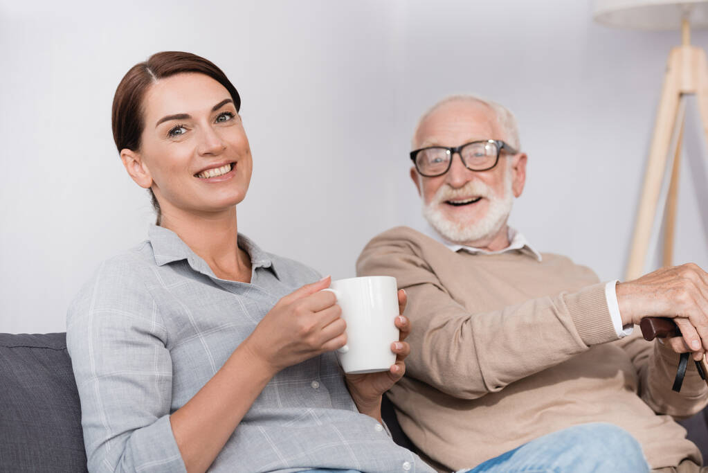 donna felice con una tazza di tè guardando la fotocamera mentre seduto vicino al padre allegro - Foto, immagini
