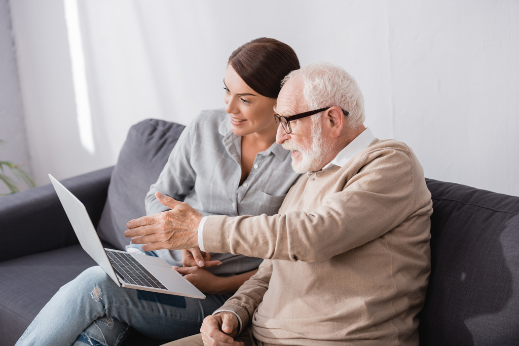 пожилой мужчина указывает рукой на ноутбук рядом с взрослой дочерью, сидя на диване дома - Фото, изображение