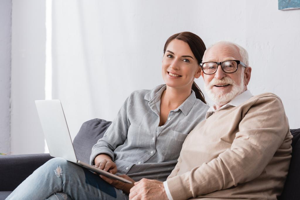 smiling brunette woman looking at camera while sitting near aged father and using laptop - Photo, Image