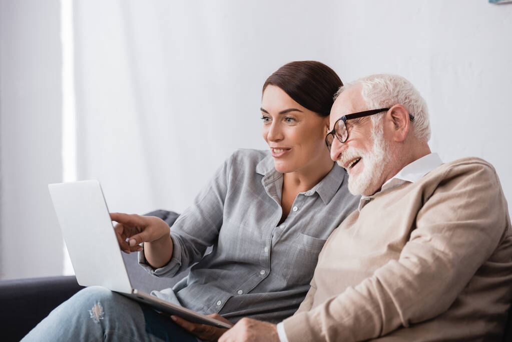 smiling woman pointing with finger at laptop near aged smiling father - Photo, Image