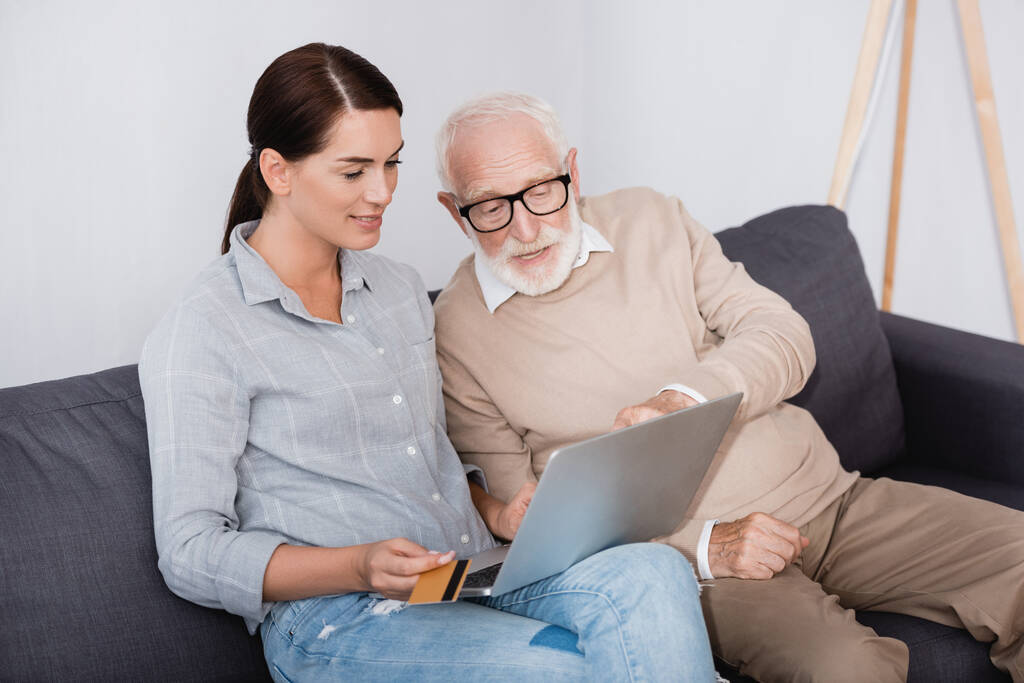 Brünette Frau hält Kreditkarte, während sie Laptop zusammen mit älterem Vater benutzt - Foto, Bild