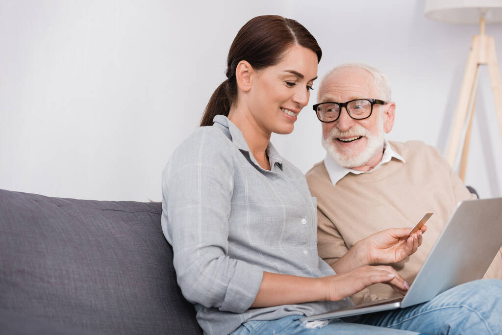 smiling woman using laptop and holding credit card near aged father at home - Photo, Image