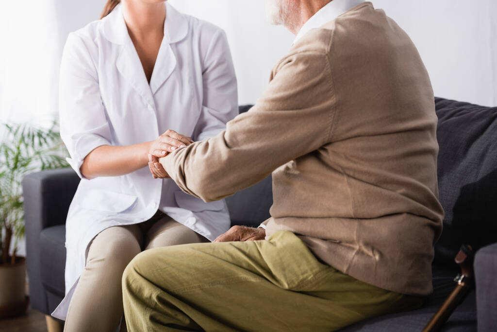 vista parcial del anciano y la enfermera geriátrica tomados de la mano mientras están sentados en el sofá  - Foto, Imagen