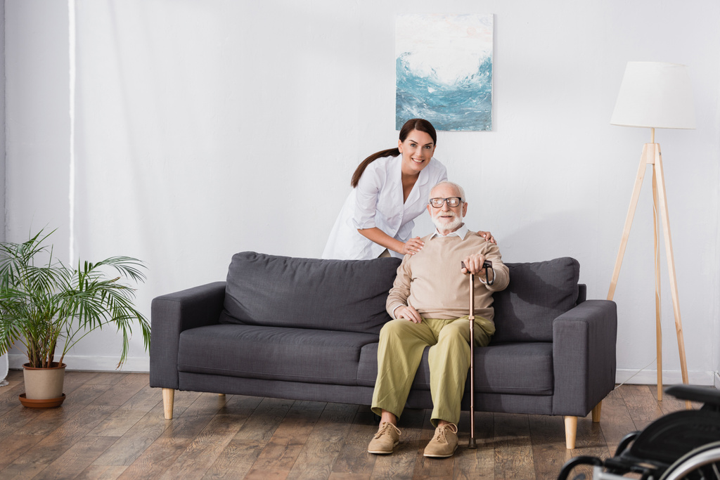 улыбающаяся медсестра обнимает пожилого мужчину сидящего на диване с тростью - Фото, изображение
