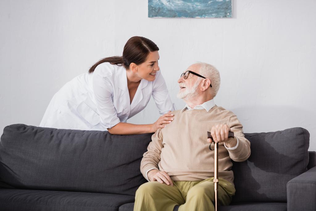 enfermera geriátrica sonriente hablando con un anciano sentado en un sofá en casa - Foto, Imagen