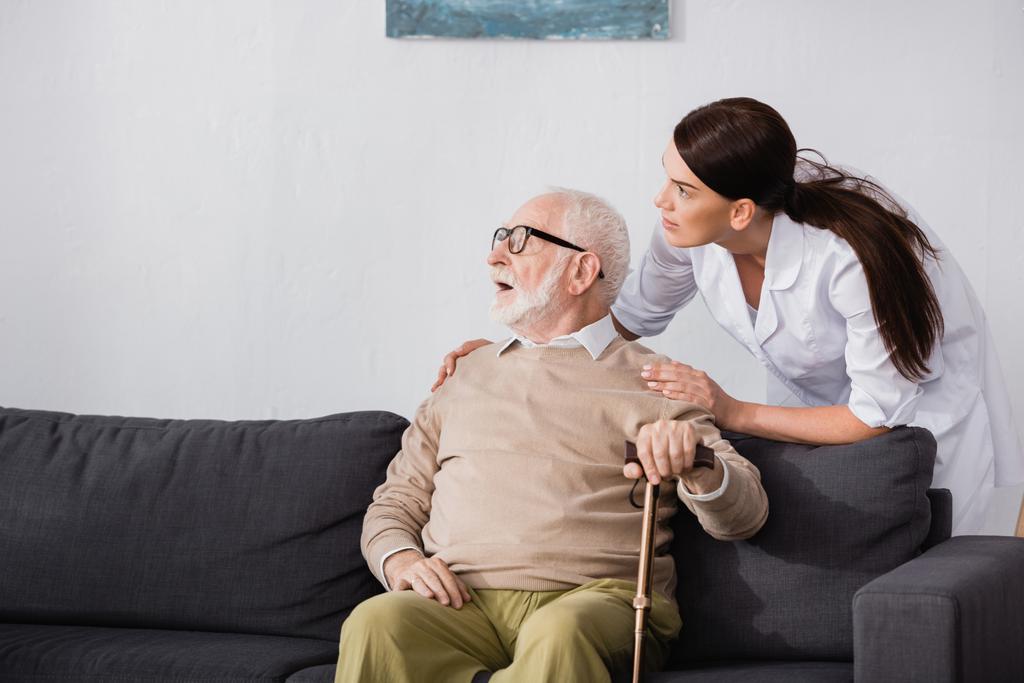 aged man looking away while sitting on sofa near social worker - Photo, Image