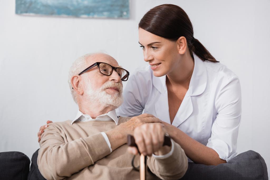 γηριατρική νοσοκόμα και ηλικιωμένος άνδρας κοιτάζονται στο σπίτι - Φωτογραφία, εικόνα