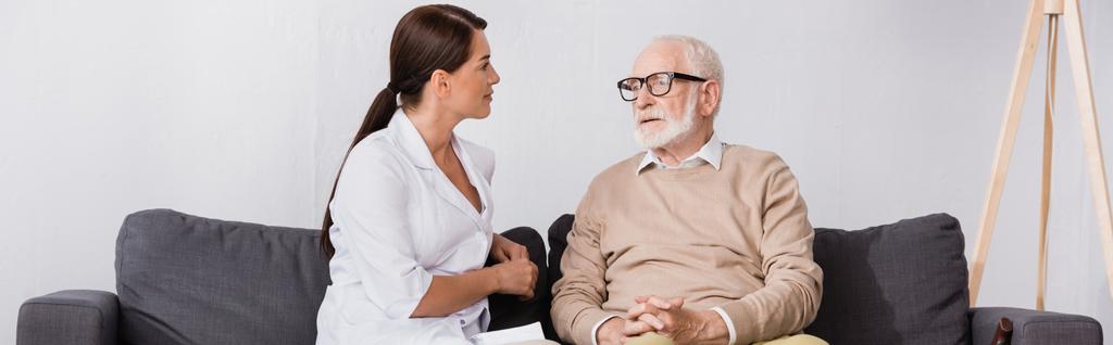 geriatrinen sairaanhoitaja puhuu ikääntyneelle miehelle istuessaan sohvalla kotona, banneri - Valokuva, kuva