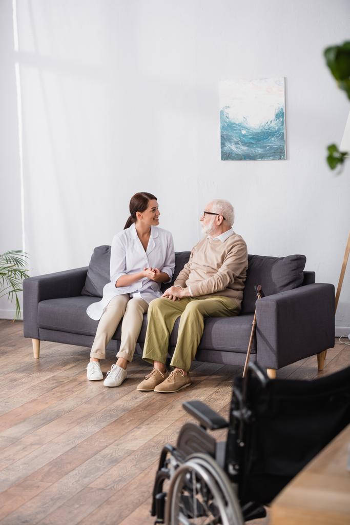 assistente social conversando com o homem idoso sentado no sofá em casa com cadeira de rodas em primeiro plano - Foto, Imagem