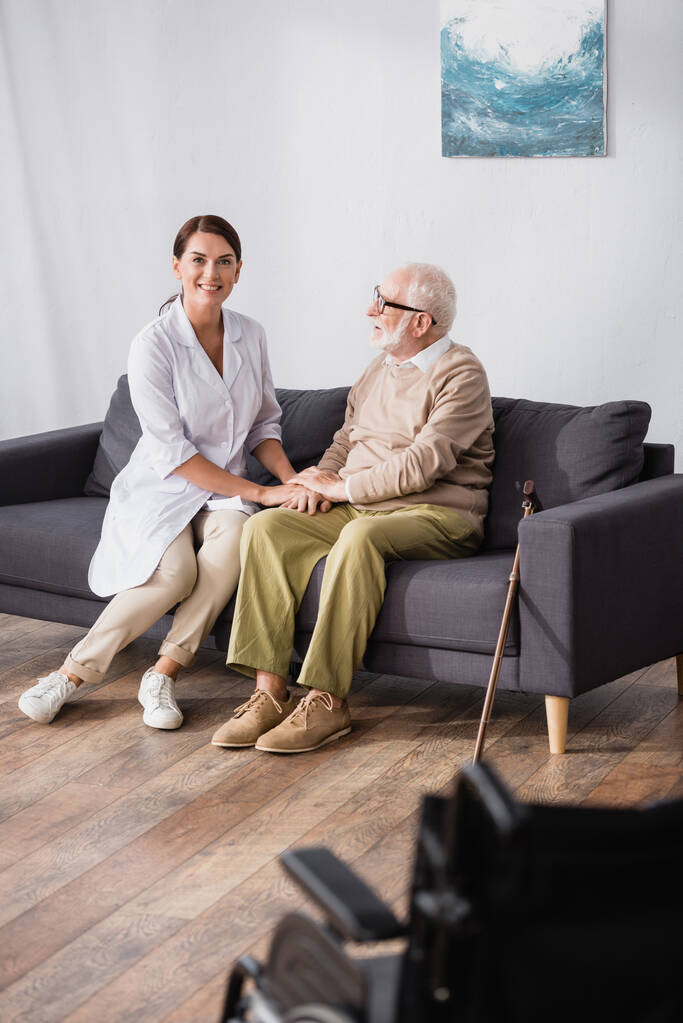 cheerful geriatric nurse holding hands with aged man sitting on sofa at home - Photo, Image
