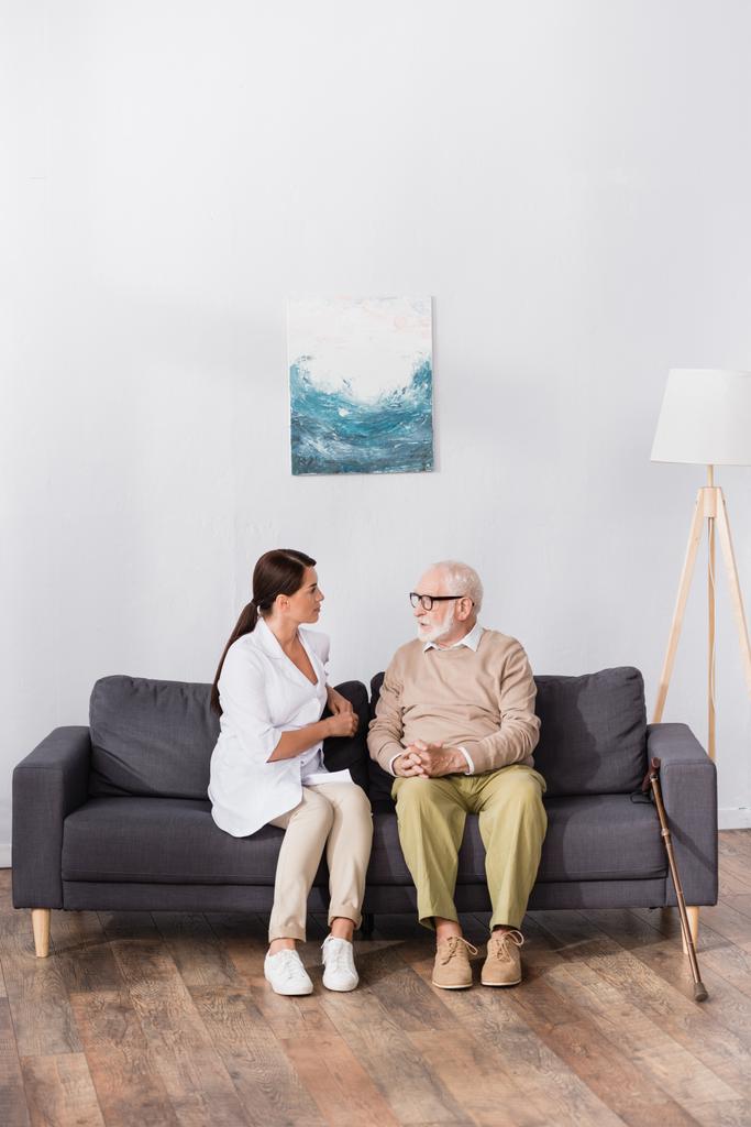 пожилой мужчина в очках разговаривает с пожилой медсестрой, сидя дома на диване  - Фото, изображение