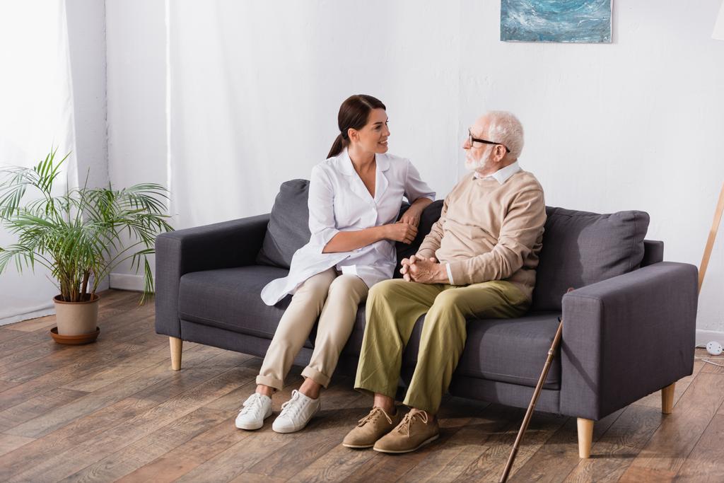 μελαχρινή κοινωνική λειτουργός και ηλικιωμένος άνδρας να μιλάει ενώ κάθεται στον καναπέ στο σπίτι - Φωτογραφία, εικόνα