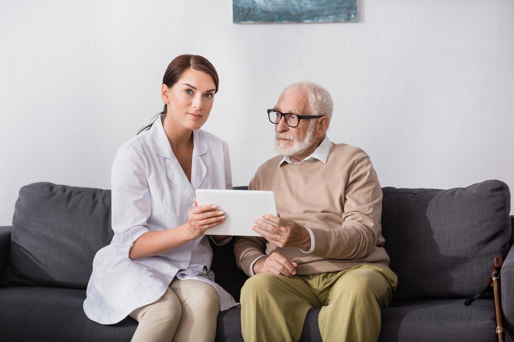 Брюнетка социальный работник показывает цифровой планшет пожилым мужчинам на диване дома - Фото, изображение